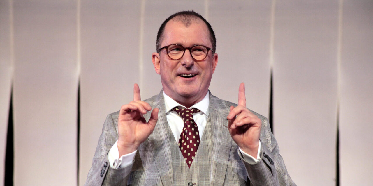 Der Schauspieler Martin Lindow steht in Anzug und Krawatte lächelnd vor einer Kulisse. Er hat die Arme angewinkelt, beide Hände sind nach oben gerichtet. Beide Finger zeigen in einer Geste, die Aufmerksamkeit erregen soll, nach oben.