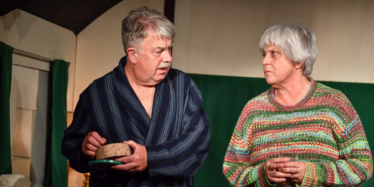 Die Schauspieler Peter E. Funck und Mona Seefried stehen vor einer Kulisse und disktueieren miteinander. Peter E. Funck hält eine geöffnete Dose in der linken Hand und sieht so aus, als ob er etwas herausnehmen möchte.