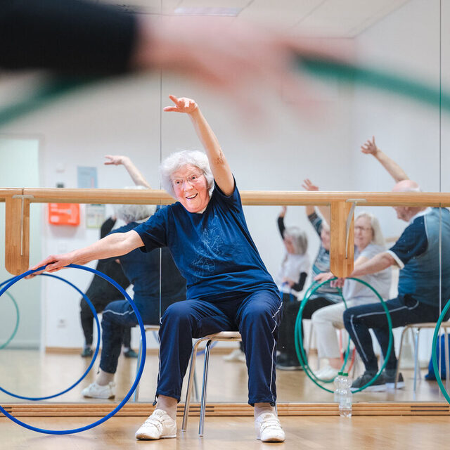 Eine ältere Frau streckt einen Arm nach oben und turnt mit einem Reifen