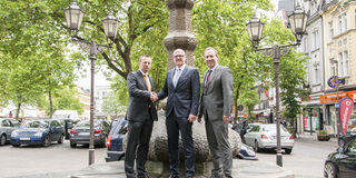Drei Männer stehen vor einem Brunnen mit einer Steinskulptur.