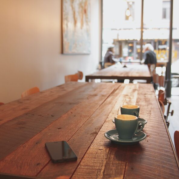Das Bild zeigt einen dunklen Holztisch in einem Café; auf dem Tisch befinden sich 2 Tassen und ein Handy