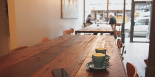 Das Bild zeigt einen dunklen Holztisch in einem Café; auf dem Tisch befinden sich 2 Tassen und ein Handy