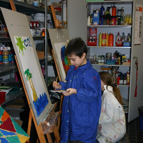 Junge mit Malkittel und Palette vor Leinwand. Er malt eine Insel im Meer.