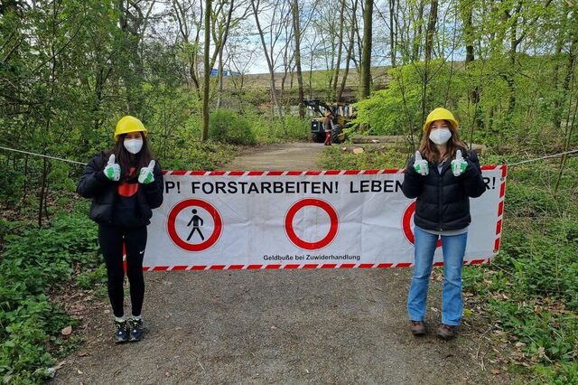 2 Mädchen stehen auf einem Waldweg, hinter Ihnen ein Banner "Stopp! Forstarbeiten! Lebensgefahr!