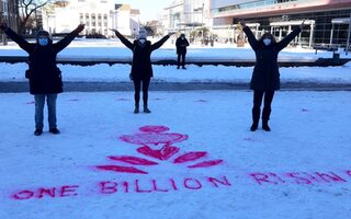 Die 3 Mitarbeiterinnen stehen mit Maske und erhobenen Armen hinter dem in den Schnee gesprühten LOGO von One Billion Rising