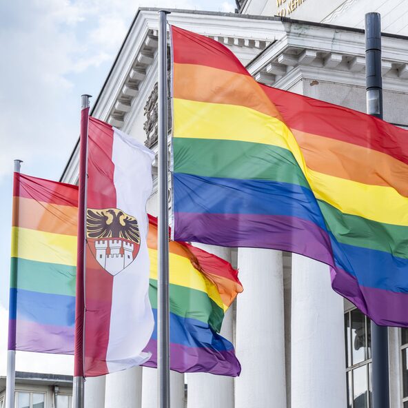 Zu sehen sind eine Flagge mit Stadtwappen und 2 Flaggen in den Farben des Regenbogens