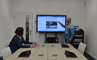 Arbeitsraum mit großem Bildschirm und PC-Stationen auf den Tischen