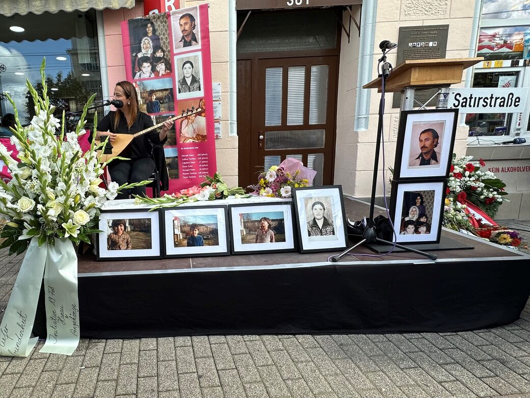 Aufbau der Bühne an der Gedenkfeier, links ein großes Trauergesteck, vorne Bilder der Opfer, dahinter die Musikerin, rechts ein Rednerpult mit einem Straßenschild auf dem steht "Satirstraße"