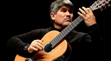 Ein Foto von Piraí Vaca vor einem schwarzen Hintergrund. Der Musiker trägt ein schwarzes Langarmshirt und spielt auf einer Akustikgitarre