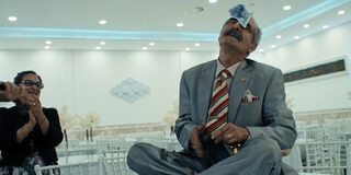 Abschnitt aus dem Film, türkischer Mann im Anzug sitzt auf einem Tisch und hat einen Geldschein auf der Stirn, an der Seite steht eine klatschende Frau