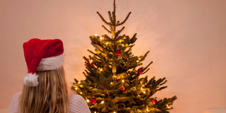 im Hintergrund ein geschmückter und leuchtender Christbaum, davor von hinten eine Frau alleine, mit einer roten Weihnachtsmütze und langen blonden Haaren, die auf den Christbaum schaut.