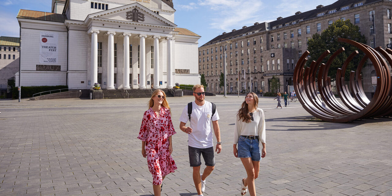 Drei Personen laufen über den König-Heinrich-Platz vor dem Stadttheater Duisburg