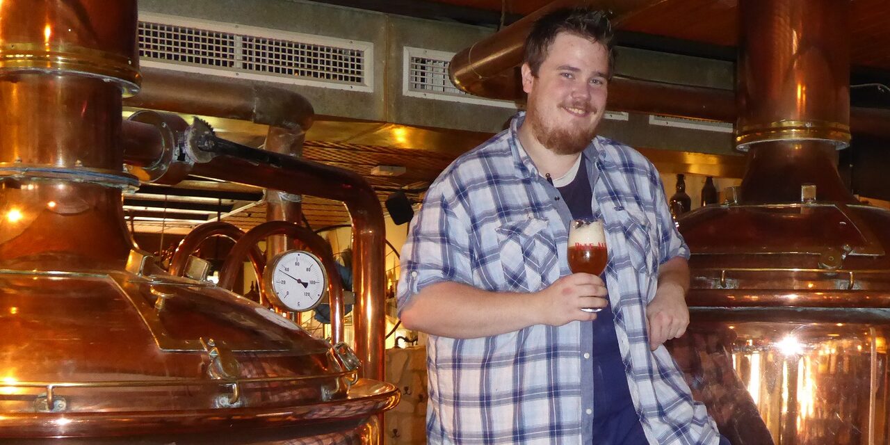 Ein Mann lehnt an einem Braukessel mit einem Glas Bier in der Hand.