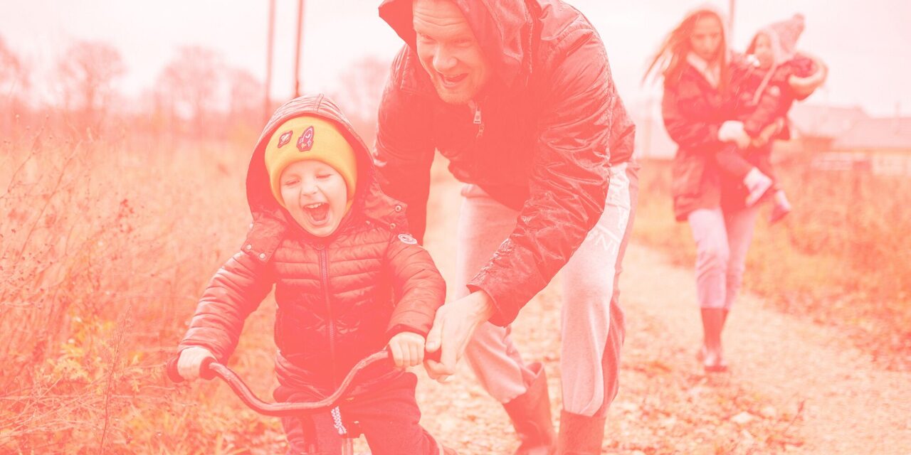 Eltern mit zwei Kinder auf einem Weg. Ein Kind fährt Fahrrad, eins wird getragen